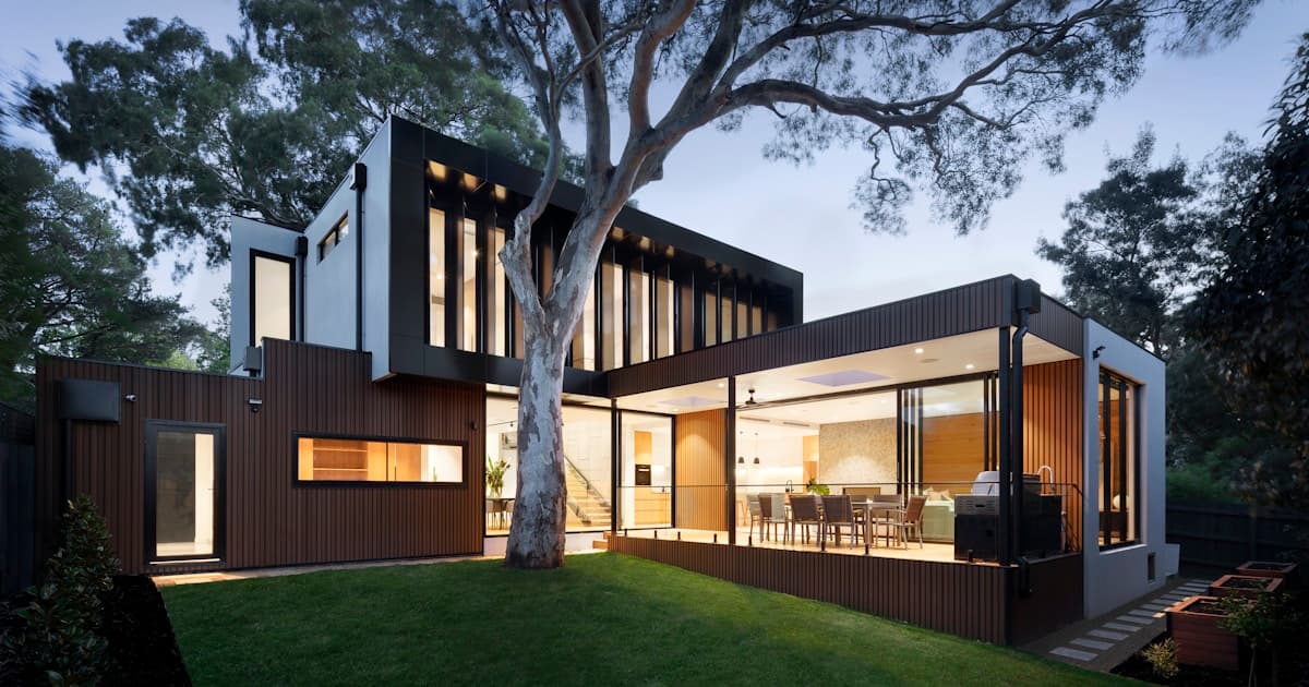 Modern two-story home with dark board-and-batten fiber cement siding, large glass windows, and covered outdoor patio at dusk